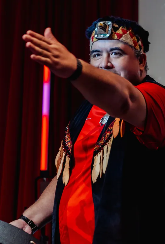 A tight portrait of a Squamish Nation performer mid-gesture, traditional headband and regalia vivid against the red curtain.