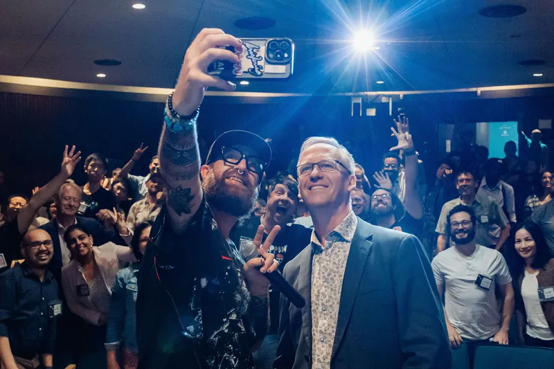 The packed hall erupts with jubilant energy behind the two figures, capturing the meetup's high-water moment.