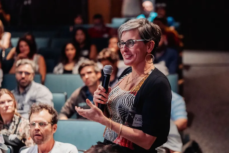 A community member in a black cardigan takes the mic and addresses the room with a broad, confident smile.