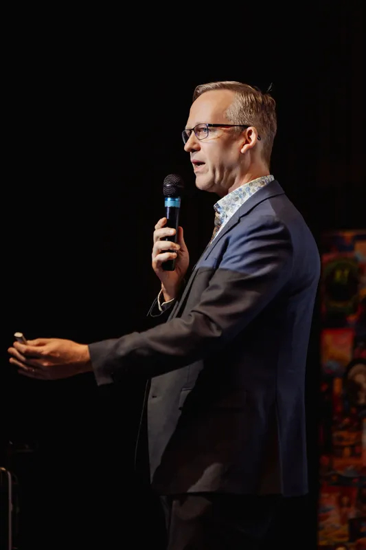 A presenter speaking into the mic extends their free arm wide with palm open in an expansive gesture. Colorful wall artwork is visible at the right edge of the frame against a dark stage.