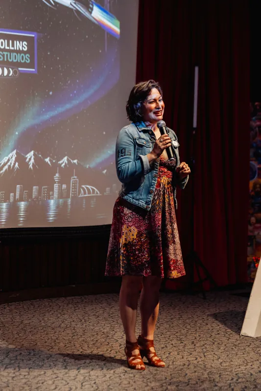 A full-length view of Mayumi Rollins commanding the stage with the Vancouver skyline graphic and aurora projection glowing behind her.