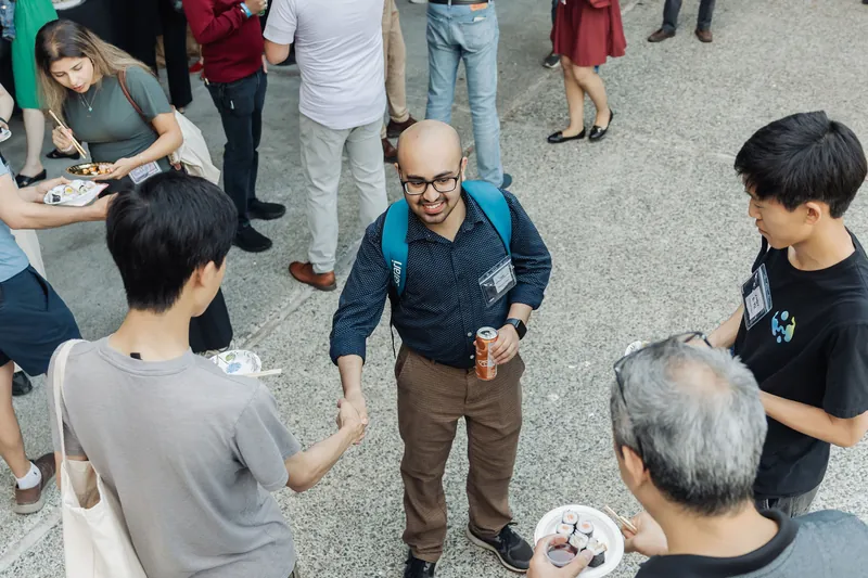 An overhead view captures an attendee reaching for a handshake on the outdoor plaza as food plates circulate among the crowd.
