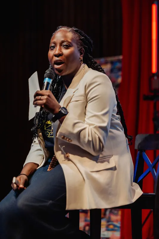 Seated on a stool with mic and notes, Anthonia Ogundele leans into her talk against a warm red-curtained backdrop.