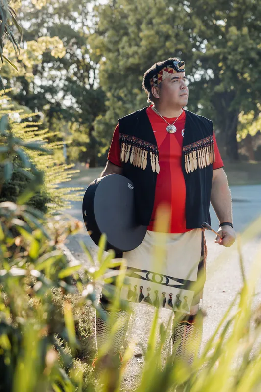 The same performer pauses on the garden path, drum at his side, gazing into the distance in warm evening light.