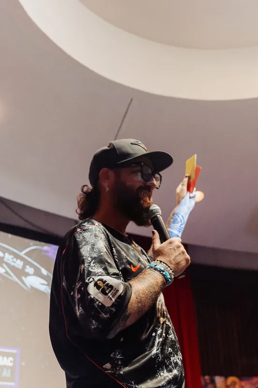A presenter in a baseball cap speaks animatedly, holding both a mic and a small slip of paper or card. The sweep of the planetarium dome curves into frame overhead.