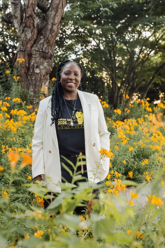 A woman in a white blazer and Ethos Lab t-shirt stands beaming amid a burst of golden blooms.