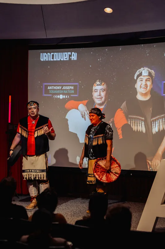 The two Squamish Nation performers stand before a large Vancouver AI projection naming them as the evening's opening act.