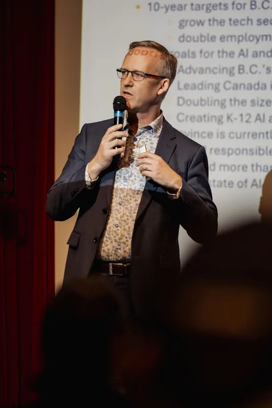 Close-up of the speaker mid-sentence, both hands raised to chest height gripping the mic, expression animated. A bright projected slide is visible behind them and a soft audience silhouette sits in the immediate foreground.