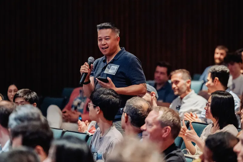 The same attendee continues his question as smiling audience members look on around him.