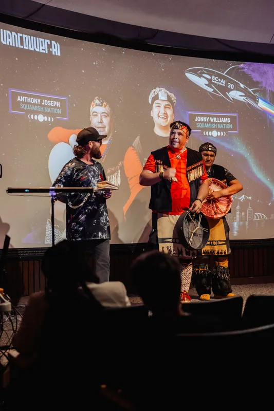 Easy laughter shared between the host and the two drum-carrying Squamish Nation performers during introductions.