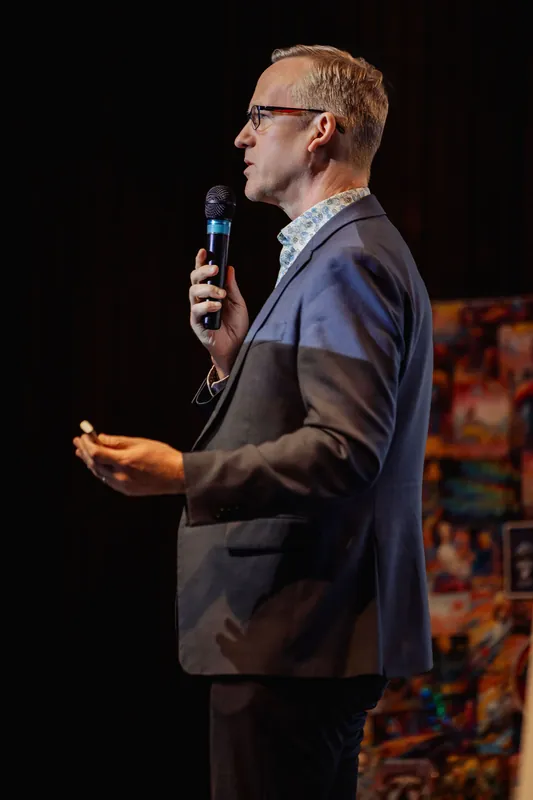 The presenter is caught mid-sentence in profile, mouth open, mic held close to the face while the free hand cups upward in a loose conversational gesture. Colorful printed artwork hangs softly out of focus in the background.
