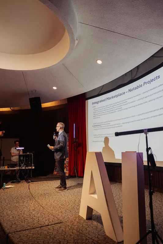 A wider stage view captures the presenter at center speaking into the mic, a projected slide glowing on the screen behind them. In the foreground, large three-dimensional white letters stand on the carpeted stage floor, and a technician is visible at a mixing desk on the left.