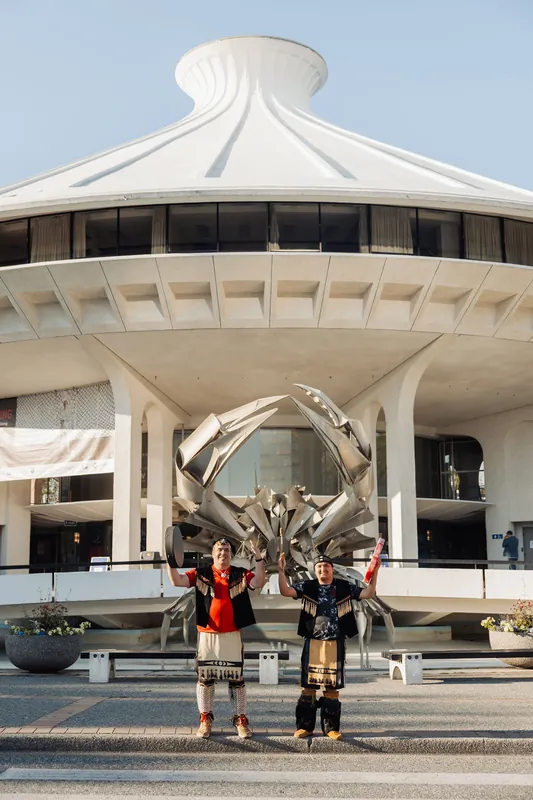The two performers raise their arms joyfully in front of the H.R. MacMillan Space Centre's crab sculpture and dome.