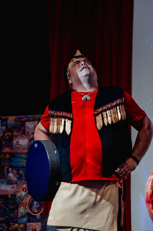 An Indigenous performer tilts his gaze upward in a moment of quiet reverence, drum resting at his side on the theatre stage.