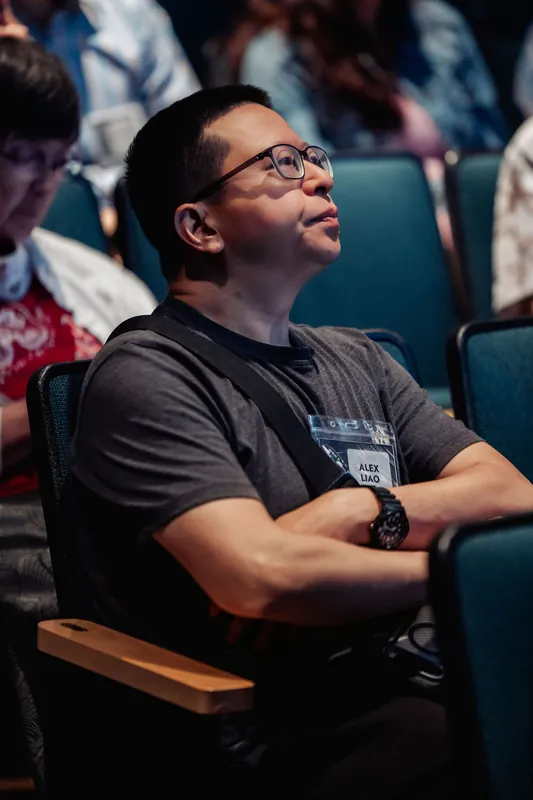 A badge-wearing attendee sits with arms folded and eyes angled upward toward the stage in quiet focus.