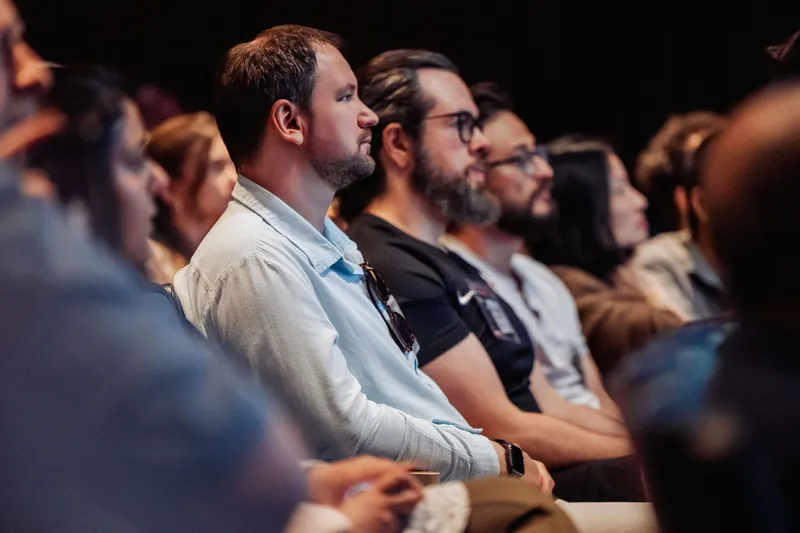 Two attendees lean forward in their seats, expressions intent, as the stage program commands the room's attention.