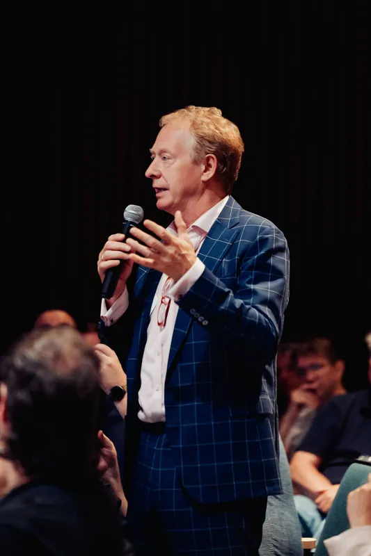 A speaker in a blue plaid blazer holds the mic and addresses the audience inside the theatre.