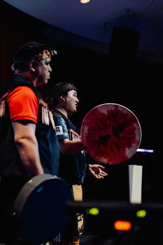 Two performers stand in close profile on stage, drums in hand, caught in dramatic stage lighting during their ceremonial set.