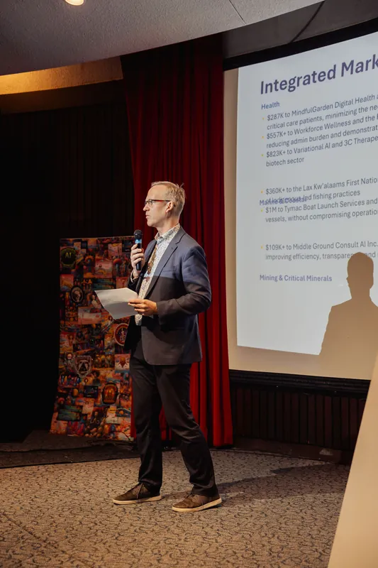 The presenter stands at the front of the room with a microphone in one hand and a sheaf of papers in the other. A large projected slide is visible behind them, and colorful artwork decorates the left wall of the venue.