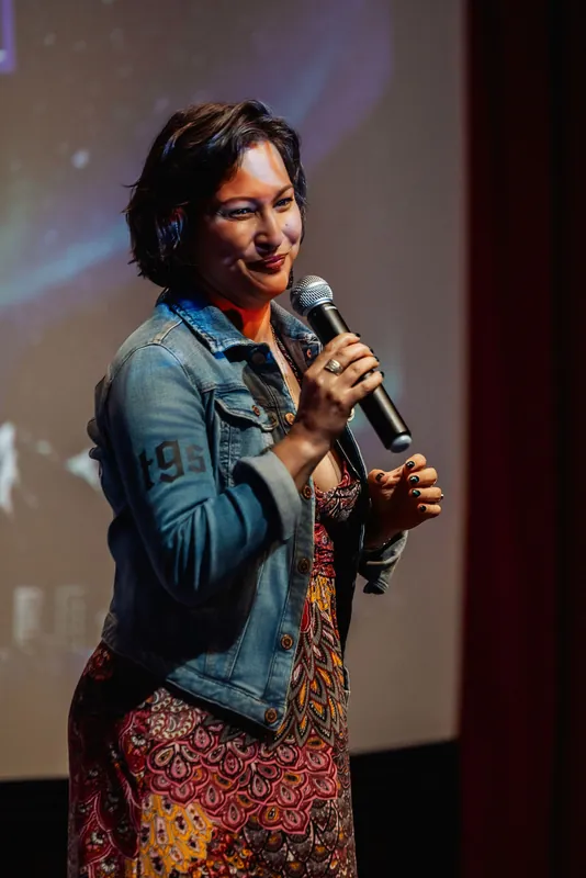 Mayumi Rollins pauses with a warm laugh during her talk, the starfield backdrop soft behind her denim jacket.