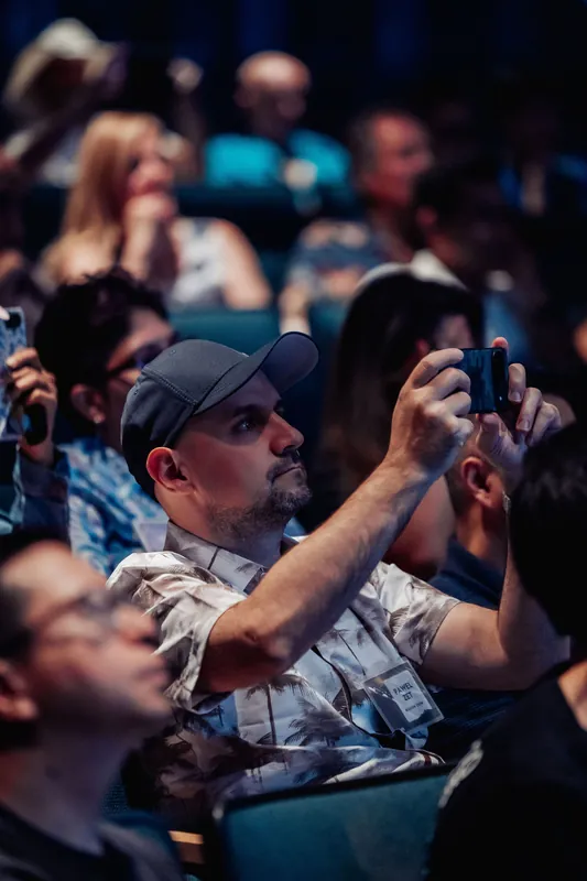 A man in a patterned shirt and cap photographs the stage from his seat as the packed planetarium audience stretches behind him.