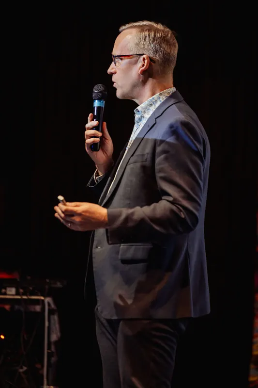A presenter in a dark blazer and patterned shirt stands in profile against a black stage backdrop, holding a handheld microphone raised near their mouth and a slide clicker in the other hand. Stage equipment is faintly visible in the lower corner.