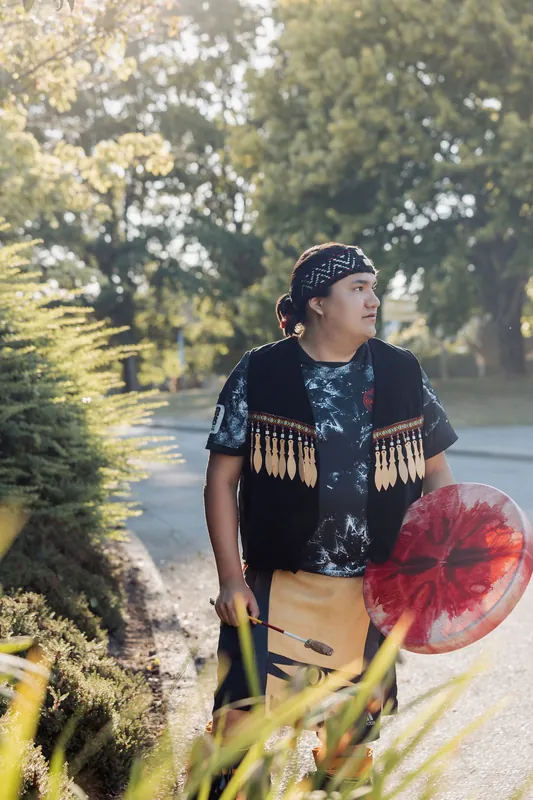 An Indigenous performer carrying a traditional hand drum walks through sun-dappled gardens in golden-hour light before the evening program.