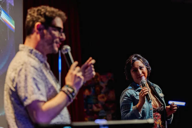 Two speakers share the stage in a candid handoff moment, Mayumi Rollins listening intently while holding her mic.