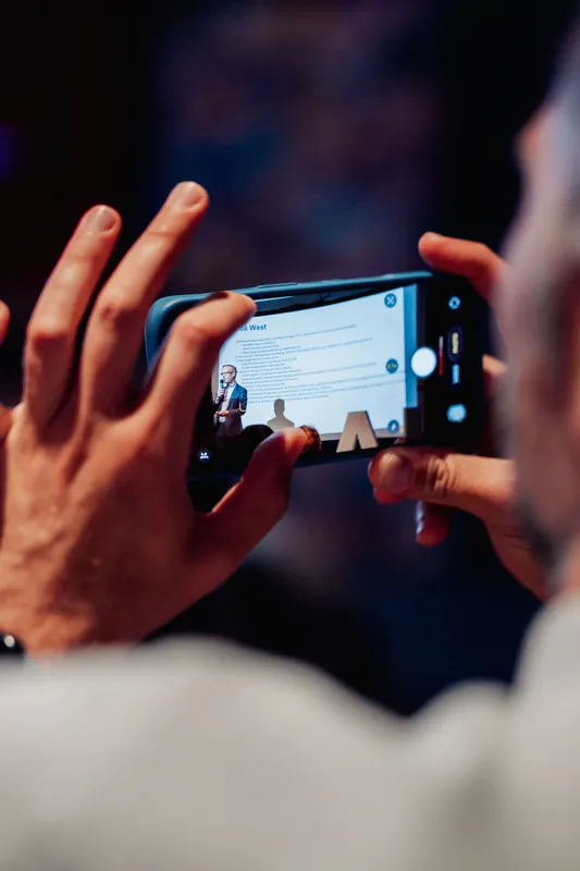 An attendee holds up a phone to capture the speaker and policy slides from their seat in the audience.