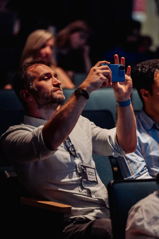 A bearded attendee raises a blue phone to film the stage while seated in the dimly lit Space Centre auditorium.