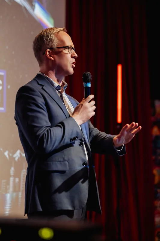 The presenter faces the audience with the microphone held at chest height, one hand extended palm-up in a conversational gesture. A projected presentation slide occupies much of the background behind them.