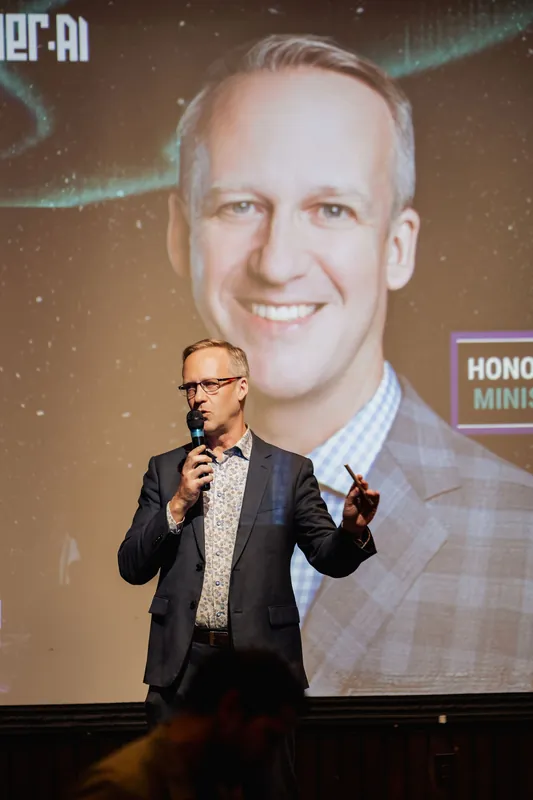 The host gestures outward with a clicker in one hand while holding the mic close to their mouth. Behind them, the projected portrait fills nearly the entire screen. A few silhouetted heads are visible at the very bottom of the frame.