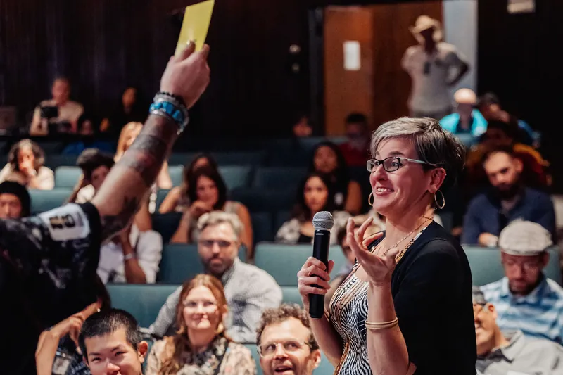 A community member rises to speak with the mic in the packed Space Centre auditorium, a yellow card lifted nearby.