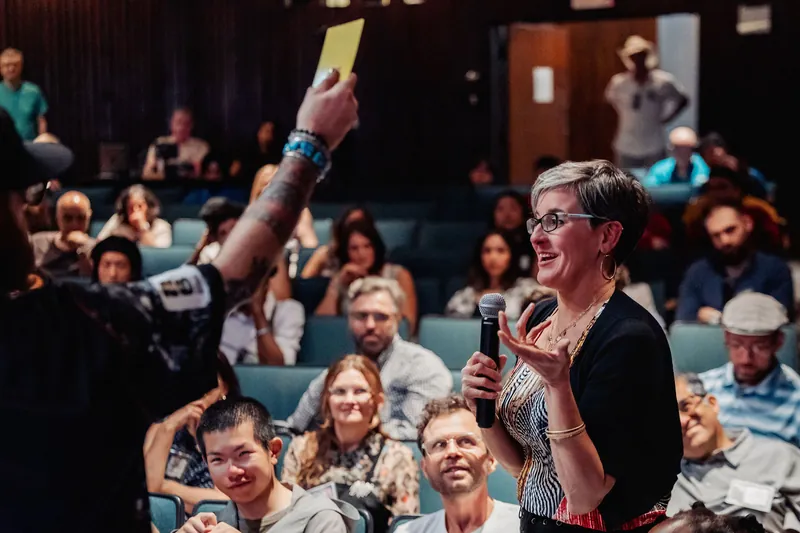 Another moment in the same exchange, the attendee speaks while the teal-seated crowd watches attentively behind her.