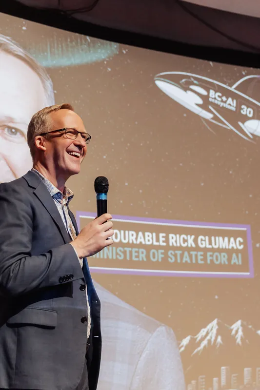The speaker laughs broadly, mic raised, in a candid moment on the lit stage. An event-branded projection fills the curved screen, and a portrait-style graphic is faintly visible behind the text overlay.