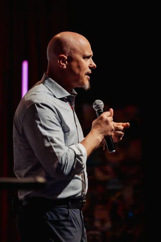 A speaker leans into his point, caught in sharp profile, microphone raised, against a softly bokeh-blurred audience.