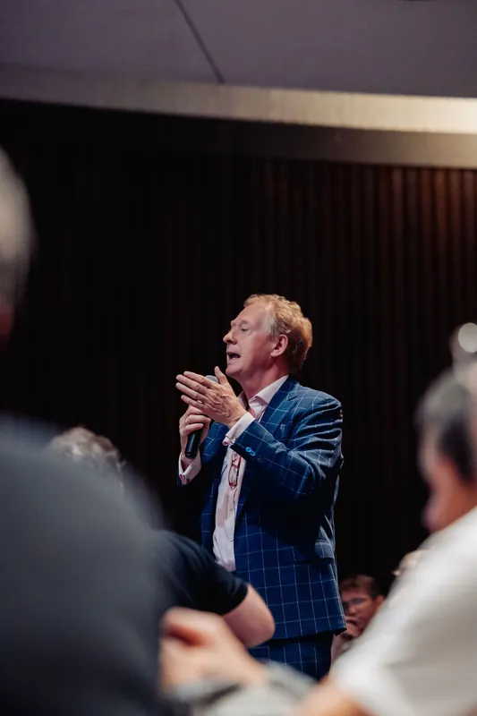 A speaker in a blue plaid blazer addresses the room with animated, expressive hands during the indoor program.