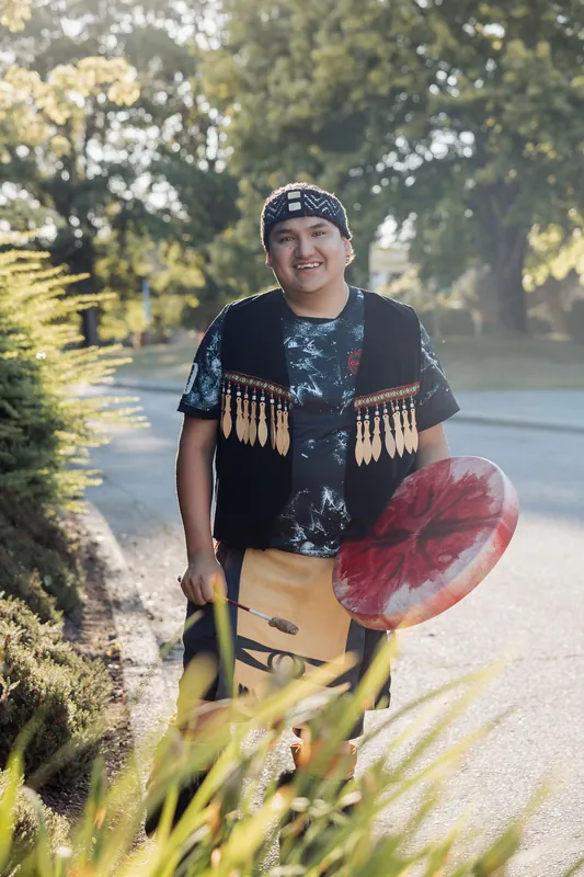 A younger performer in traditional regalia smiles broadly while carrying a red-painted drum along the golden-hour grounds path.