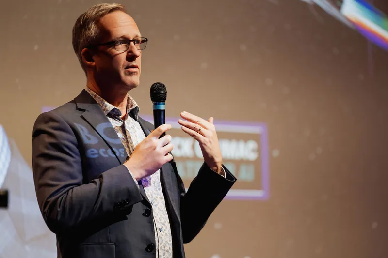 A tighter frame catches the presenter with both hands raised — mic in one, the other lifted with fingers loosely spread in a conversational gesture. A projected slide fills the warm-toned background.