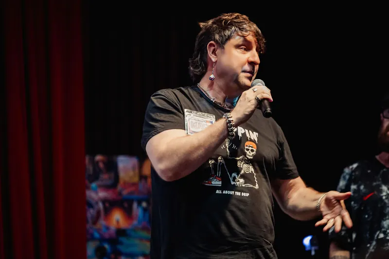 A speaker in a black graphic tee takes the microphone, his confident expression lit close-up against a dark stage backdrop.