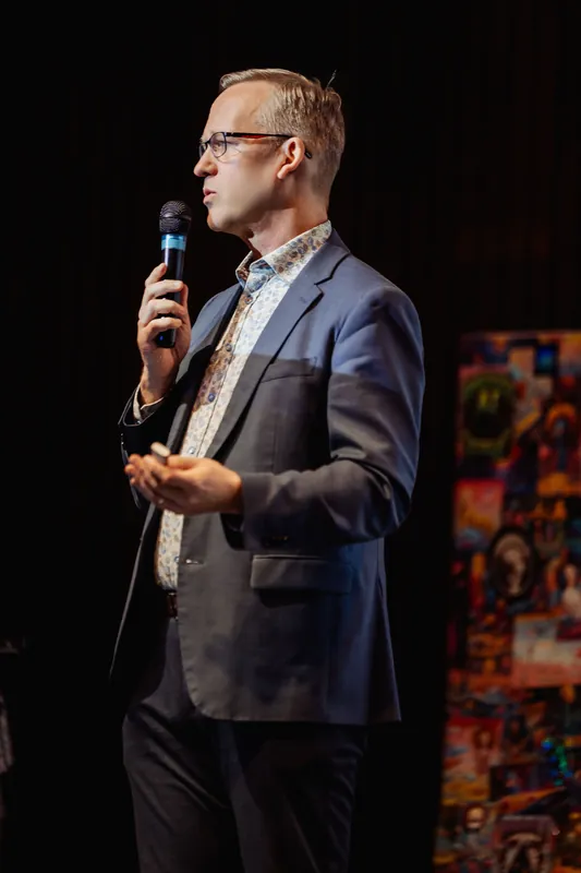 Three-quarter profile of the speaker in the dark stage setting, mic raised and free hand relaxed. Vivid colorful banners or artwork panels are visible to their right, contrasting with the dark surround of the stage.