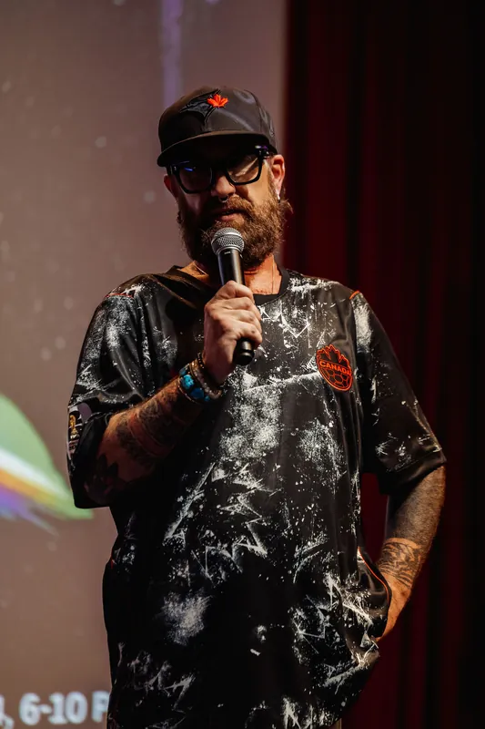 A speaker in a tie-dye shirt and dark cap holds the mic under the warm glow of a planetary projection inside the Space Centre.