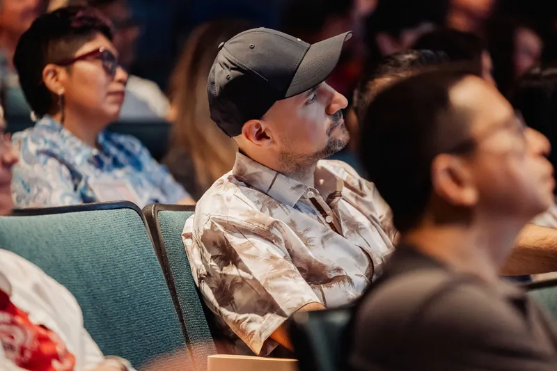 A cluster of attendees including a man in a cap and floral shirt all look upward toward the stage during the indoor program.