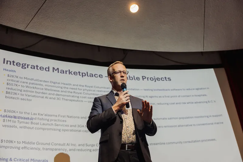 Wider stage view with the speaker mid-gesture, one hand raised and the other holding the mic. The curved planetarium dome and a single ceiling spotlight frame the shot from above, with the full projection screen visible beneath.