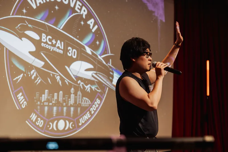 A performer at the mic gestures hand-raised before the Vancouver AI Meetup's Mission 30 badge projection at the Space Centre.