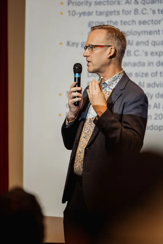 Pure profile shot of the speaker from the left: mic raised in the right hand, left hand lifted in an open-palm gesture near the chest. A bright projected slide fills the background. Audience silhouettes are just visible at the bottom.