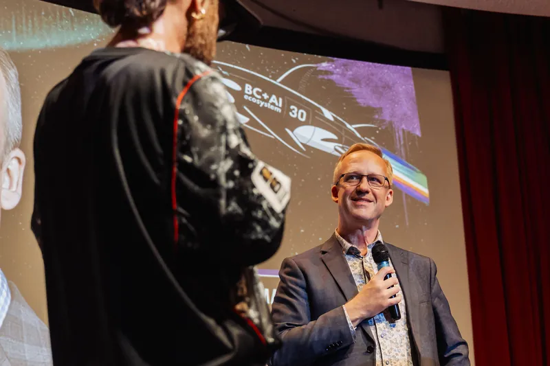 Shot looking past one presenter's shoulder toward the second speaker, who smiles broadly while holding a mic. An event-themed graphic is projected on the curved screen in the background.