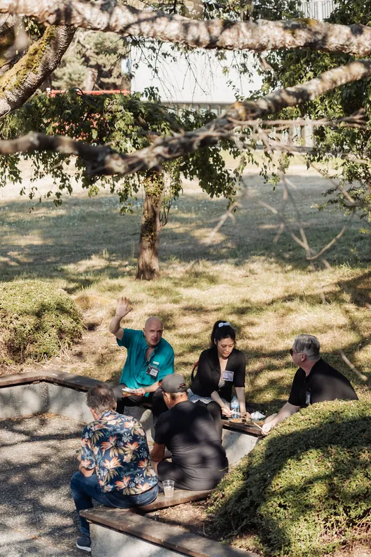 A small group gathers around a low stone table on the Space Centre grounds, one attendee gesturing mid-story beneath the tree canopy.