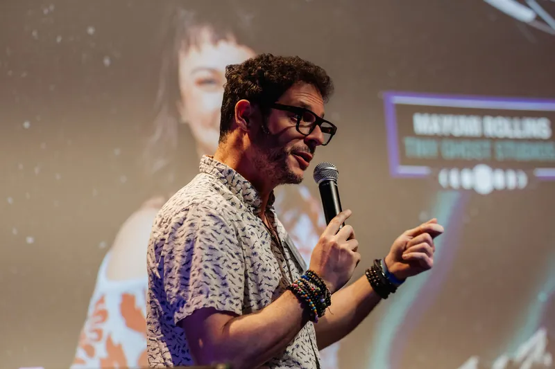 The event host speaks into a mic with the Mayumi Rollins speaker slide still glowing on the projection screen behind him.