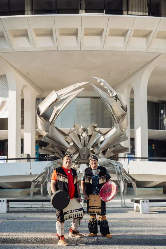 Both performers hold their drums and look toward the camera with the full Space Centre facade filling the background.